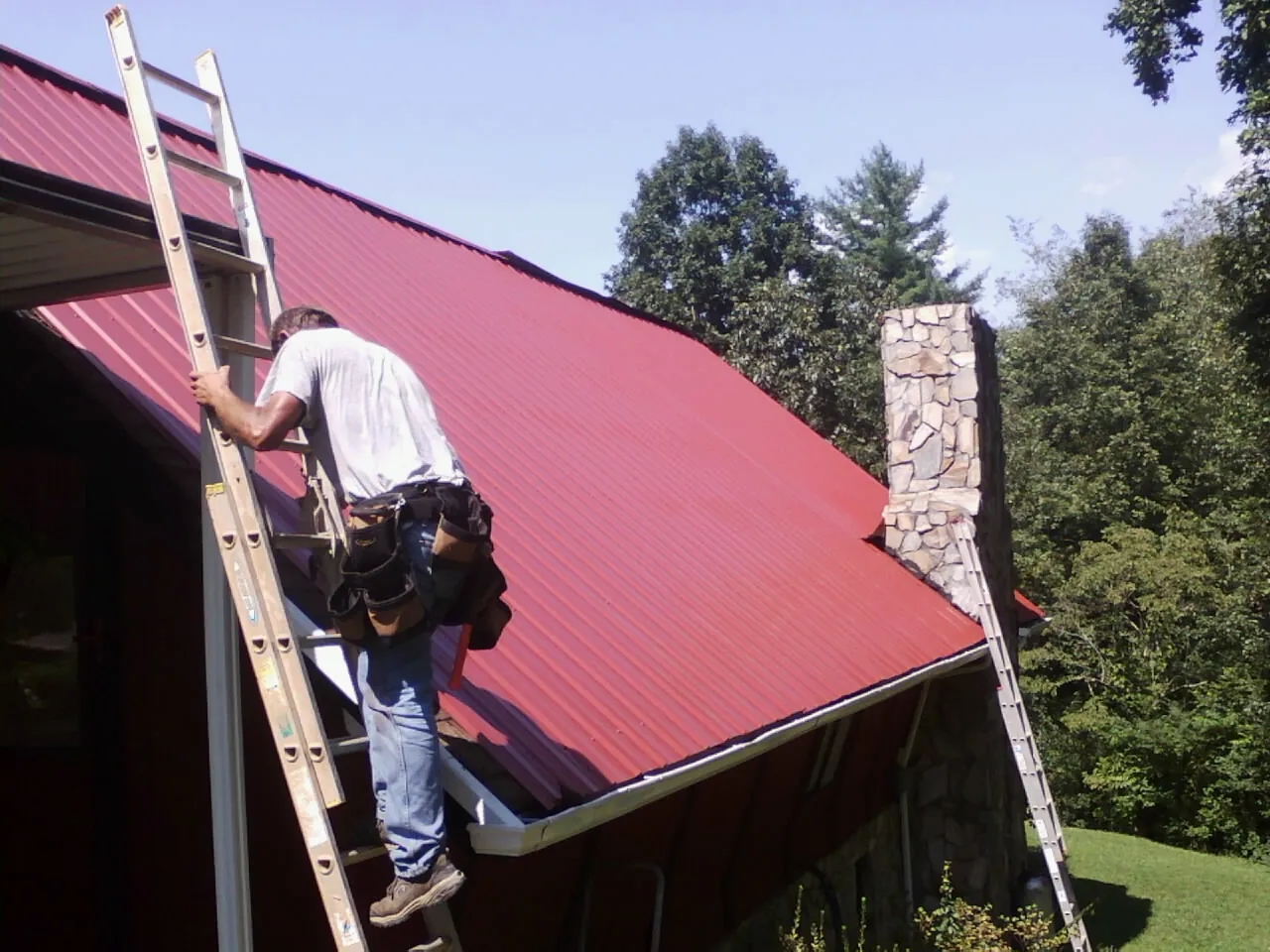 Roofer on a ladder beside a red metal roof and stone chimney — Soffit & Fascia Repair by Roofing Brothers in Kimball